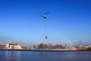 Fototapeta na wymiar Helicopter KA-32 MES draws water from the lake to extinguish a forest fire. In the background is a blue sky.