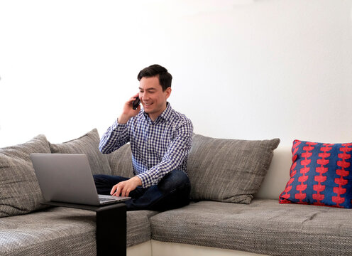 Smiling Man Working From Home Typing On Laptop And Making A Phone Call