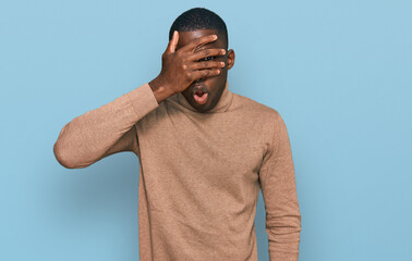 Young african american man wearing casual winter sweater peeking in shock covering face and eyes with hand, looking through fingers with embarrassed expression.