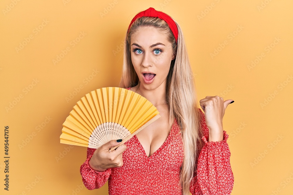 Poster Young caucasian woman waving hand fan cooling air in summer pointing thumb up to the side smiling happy with open mouth