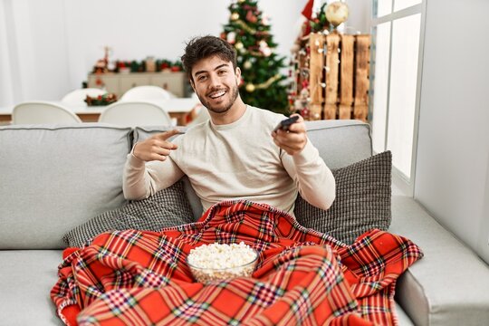 Young hispanic man eating popcorn sitting on sofa by christmas tree smiling happy pointing with hand and finger
