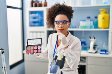 Young african american woman working at scientist laboratory doing make up serious face thinking about question with hand on chin, thoughtful about confusing idea