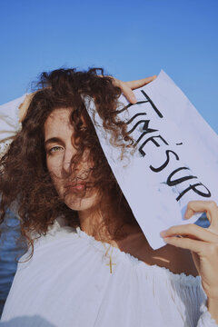 Young woman in light dress holding a handwritten paper with "Time's Up" at the beach