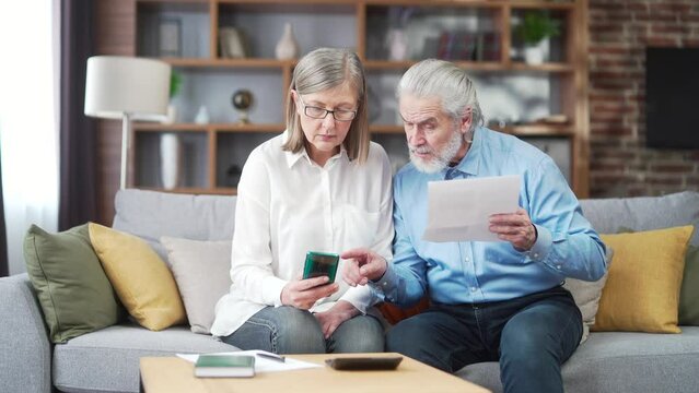 senior family couple, gray haired old man and woman pay for services enter data documents using application mobile smartphone. Retirees at home. utility bills, checks use computer smart phone