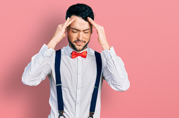 Hispanic man with beard wearing hipster look with bow tie and suspenders with hand on head for pain in head because stress. suffering migraine.
