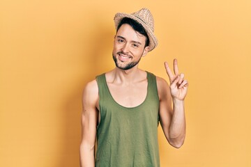 Young hispanic man wearing summer hat smiling with happy face winking at the camera doing victory sign. number two.