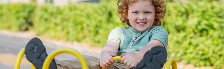 excited redhead kid looking at camera while riding seesaw in park, banner.