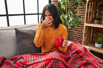 Young hispanic woman sitting on the sofa drinking a coffee at home smelling something stinky and disgusting, intolerable smell, holding breath with fingers on nose. bad smell