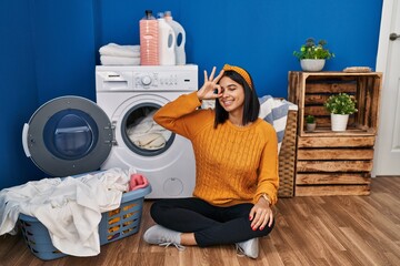 Young hispanic woman doing laundry doing ok gesture with hand smiling, eye looking through fingers with happy face.