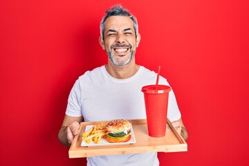 Handsome middle age man with grey hair eating a tasty classic burger with fries and soda smiling and laughing hard out loud because funny crazy joke.