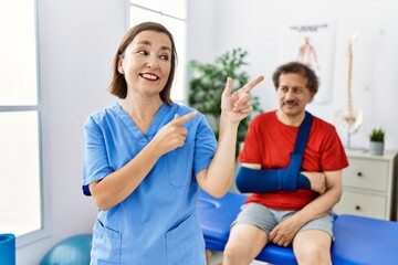 Obraz premium Middle age doctor woman with patient with arm injury at rehabilitation clinic smiling and looking at the camera pointing with two hands and fingers to the side.