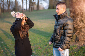 Caucasian couple in casual wear is in the spring park. Happy and smiling they joke among themselves: he pretends to puddle her and with a quick movement of her he lowers her cap covering her face.