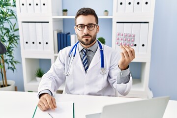 Handsome hispanic man wearing doctor uniform holding prescription pills thinking attitude and sober expression looking self confident