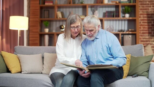 A lovely senior couple watching Memoirs photo album on sofa at home. Elderly family grandparents sitting together looking and discussing pleasant moments turnes pages with photographs, nostalgia