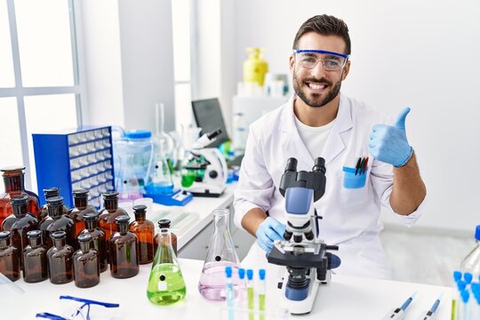 Handsome hispanic man working at scientist laboratory smiling happy and positive, thumb up doing excellent and approval sign