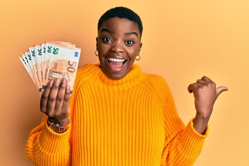 Young african american woman holding 50 euro banknotes pointing thumb up to the side smiling happy...