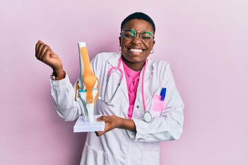 Young african american doctor woman holding anatomical model of knee joint screaming proud,...