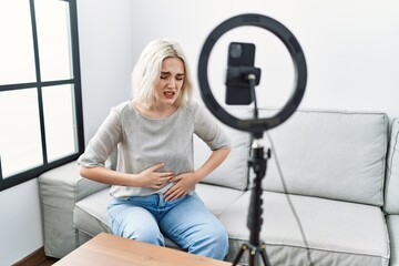 Young caucasian woman recording vlog tutorial with smartphone at home with hand on stomach because...