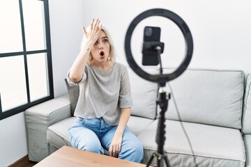 Young caucasian woman recording vlog tutorial with smartphone at home surprised with hand on head...