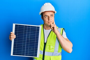 Handsome young man holding photovoltaic solar panel serious face thinking about question with hand...