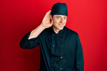 Fototapeta na wymiar Handsome young man wearing professional cook uniform and hat smiling with hand over ear listening an hearing to rumor or gossip. deafness concept.