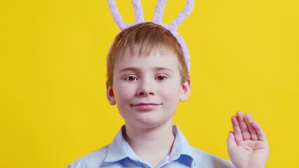 Close-up of a happy boy with rabbit ears on a yellow background. The boy waves his hand and looks at the camera. Easter