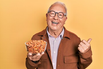 Senior man with grey hair holding fried peanuts pointing thumb up to the side smiling happy with open mouth