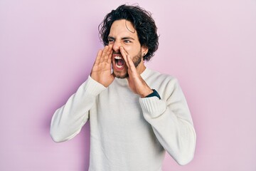 Handsome hispanic man wearing casual white sweater shouting angry out loud with hands over mouth