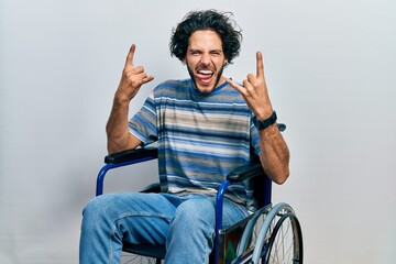 Handsome hispanic man sitting on wheelchair shouting with crazy expression doing rock symbol with...