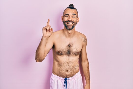 Young hispanic man shirtless wearing swimsuit pointing finger up with successful idea. exited and happy. number one.