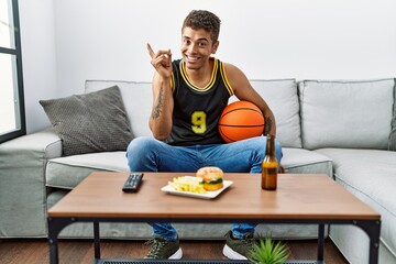 Young handsome hispanic man holding basketball ball cheering tv game smiling happy pointing with hand and finger to the side