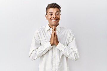 Young handsome hispanic man standing over isolated background praying with hands together asking for forgiveness smiling confident.