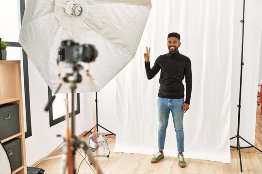 Young hispanic man with beard posing as model at photography studio showing and pointing up with fingers number two while smiling confident and happy.