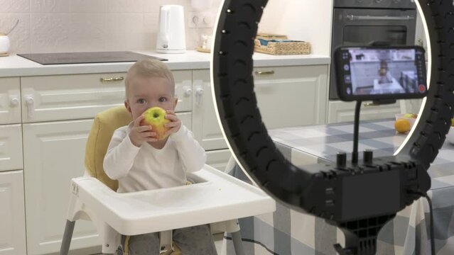 Parents record video on smartphone of kid in high chair at home, recording with mobile phone and ring light photo video of one-year-old baby boy with fruit. High quality 4k footage