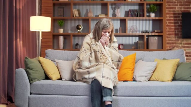 Gray hair aged elderly woman with flu symptoms sitting on couch blowing nose using napkin. sick senior female at home coughs. Use a mobile phone, call a doctor for help or advice. 