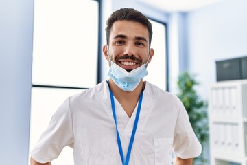Young hispanic man wearing physiotherapist uniform and medical mask at clinic