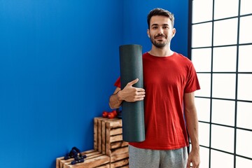 Young hispanic man wearing sportswear holding yoga mat at sport center