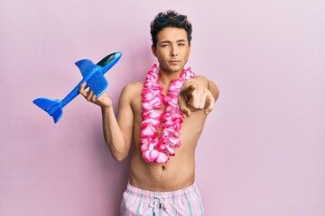 Young handsome man wearing swimsuit and hawaiian lei holding airplane toy pointing with finger to...