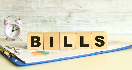 Wooden cubes lie on a folder with financial charts on a gray background. The cubes make up the word BILLS.