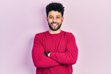 Young arab man with beard wearing casual pink sweater happy face smiling with crossed arms looking...