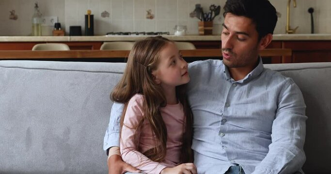 Young stepfather talks to little daughter seated on sofa in domestic room. Little girl having pleasant reliable conversation to loving dad at home. Fatherhood, kid upbringing, give wise advice concept