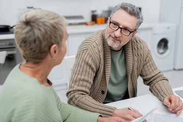 Mature man looking at blurred wife near documents in kitchen.