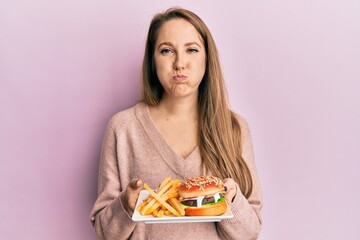 Young blonde woman eating a tasty classic burger with fries puffing cheeks with funny face. mouth inflated with air, catching air.