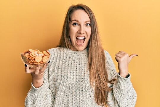 Young blonde woman holding salty biscuits bowl pointing thumb up to the side smiling happy with open mouth