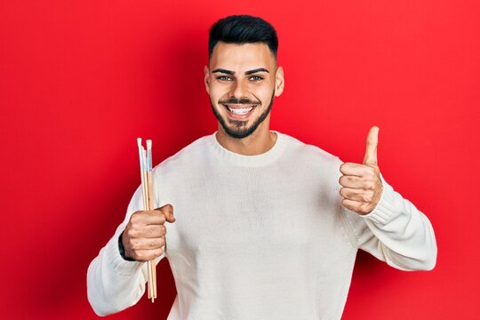 Young hispanic man with beard holding paintbrushes smiling happy and positive, thumb up doing excellent and approval sign