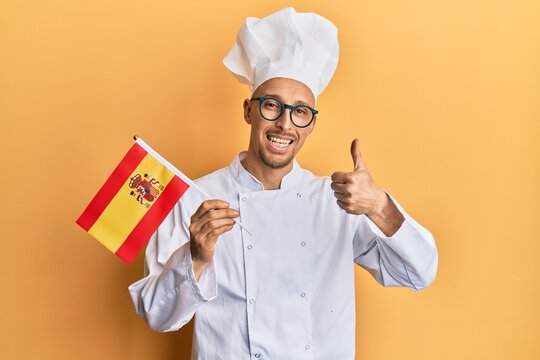 Bald man with beard wearing professional cook apron holding spanish flag smiling happy and positive, thumb up doing excellent and approval sign