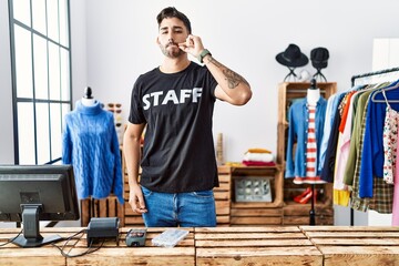 Young hispanic man working at retail boutique mouth and lips shut as zip with fingers. secret and silent, taboo talking - obrazy, fototapety, plakaty
