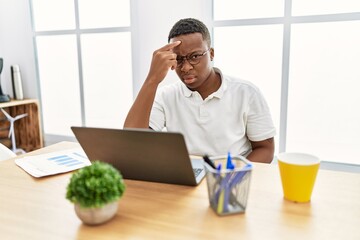 Young african man working at the office using computer laptop pointing unhappy to pimple on forehead, ugly infection of blackhead. acne and skin problem