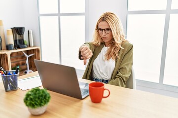 Beautiful blonde woman working at the office with laptop looking unhappy and angry showing rejection and negative with thumbs down gesture. bad expression.