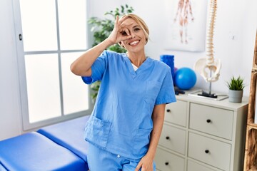 Beautiful blonde physiotherapist woman working at pain recovery clinic doing ok gesture with hand smiling, eye looking through fingers with happy face.